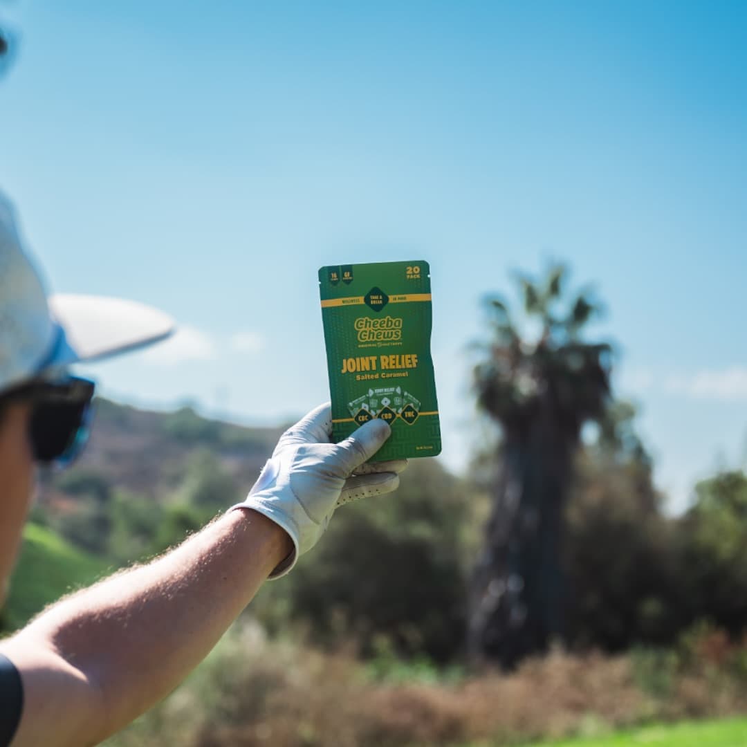 Golf gloved hand holding up a Cheeba Chews Joint Relief Salted Caramel pouch against blue sky